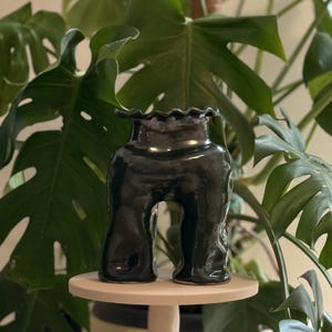 Pode incluir: Um vaso de cerâmica cinza-carvão escuro com acabamento brilhante, apresentando uma base em forma de arco distinta com duas pernas e uma borda superior ondulada e franzida. O objeto tem uma superfície lisa, reflexiva e irregular.