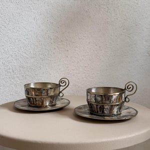 Könnte beinhalten: Zwei kleine Tassen mit passenden Untertassen, die einen Metallrahmen mit eingelegten, irisierenden Abalone-Muschelstücken aufweisen. Jede Tasse hat einen markanten, silberfarbenen Metallgriff in Form einer Spirale als Verzierung.