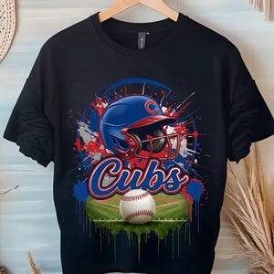 Puede incluir: Una camiseta negra de manga corta con un estampado gráfico de un casco de béisbol azul con visera roja y la letra C, la palabra Cubs en letra azul y roja, una pelota de béisbol blanca y un campo de béisbol verde sobre un fondo de salpicaduras.