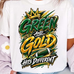 May include: A white t-shirt featuring a graphic design with the text "GREEN AND GOLD HITS DIFFERENT". The design includes a yellow crown, green and gold paint drip effects, and a football illustration with green and white details.
