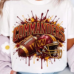 Pode incluir: Uma t-shirt branca com uma estampa gráfica exibindo a palavra COMMANDERS em letras grandes bordô e douradas. Abaixo do texto, há uma bola de futebol americano marrom e um capacete bordô com um logotipo W dourado, cercados por respingos de tinta.