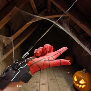May include: A red and black web-shooter wrist device featuring a metallic spring and a thin wire, worn on a hand covered by a red glove with a black web pattern. The device is constructed from black plastic components with white line accents.