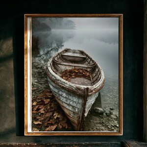 May include: A weathered wooden rowboat rests on a pebbled shore beside a calm, misty lake. The boat features peeling white paint, a wooden bench, and a collection of brown fallen leaves inside. The scene is framed in a simple wooden border.