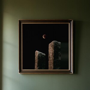 May include: A square framed wall art piece featuring a dark night sky with a glowing red lunar eclipse positioned above two tall, textured, weathered stone monoliths. The frame is dark brown with a thin gold inner border detail.