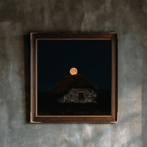 May include: A square framed wall art piece featuring a photograph of a stone cottage with a thatched roof under a dark night sky. A large, glowing orange full moon is positioned directly above the peak of the roof in the center of the frame.