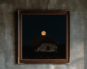 May include: A square framed wall art piece featuring a photograph of a stone cottage with a thatched roof under a dark night sky. A large, glowing orange full moon is positioned directly above the peak of the roof in the center of the frame.
