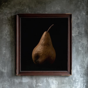 May include: A framed wall art piece featuring a single brown, textured pear centered against a solid black background. The artwork is enclosed in a dark brown wooden frame with a beaded inner edge, creating a stark contrast between the fruit and frame.