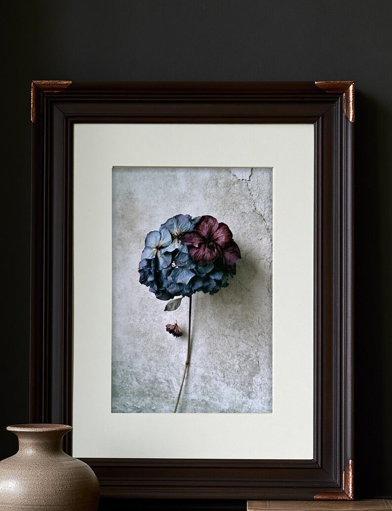 May include: A framed print featuring a single hydrangea flower with blue and deep purple petals on a long stem. The artwork is set within a dark brown wooden frame with copper-toned corner accents and a wide, off-white mat board border.