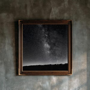 May include: A square wall art piece featuring a black and white photograph of a starry night sky above a dark, jagged horizon line. The image is encased in a dark brown, ornate wooden frame with a textured, beaded inner edge detail.