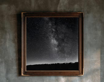 May include: A square wall art piece featuring a black and white photograph of a starry night sky above a dark, jagged horizon line. The image is encased in a dark brown, ornate wooden frame with a textured, beaded inner edge detail.