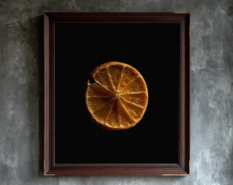 May include: A framed wall art piece featuring a single dried orange slice centered on a solid black background. The frame is a dark brown wood with a beaded inner edge detail. The orange slice displays a translucent, golden-amber texture.