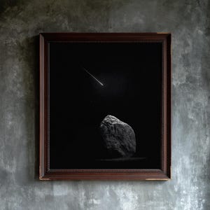 May include: A framed wall art piece featuring a dark, black background with a single bright shooting star streak in the upper portion and a large, textured, gray rock formation positioned in the lower center, all enclosed in a dark brown frame.