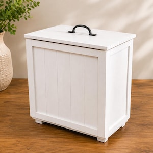 Pode incluir: Um recipiente de armazenamento retangular de madeira branca com um design de tábuas verticais no painel frontal. O recipiente possui uma tampa plana com uma alça arqueada de metal preto centralizada no topo. A estrutura é apoiada por quatro pés.