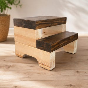 Op de afbeelding: Een houten opstapje met twee treden, voorzien van een basis van lichtgekleurd natuurlijk hout en donkerbruin gebeitste treden. Het krukje is geconstrueerd met dikke, massieve houten planken en heeft een stevig frame met boogvormige uitsparingen.