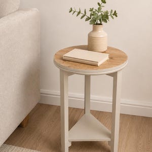 May include: A round side table with a light wood grain top and a cream-coloured frame featuring three straight legs and a triangular lower shelf. A cream-coloured vase with green eucalyptus stems and a closed book rest on the table surface.
