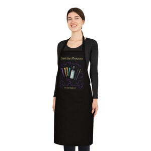 Può includere: Un grembiule nero con il testo dorato Trust the Process e un'illustrazione grafica di fermagli per capelli colorati, uno spruzzino e un uncinetto circondati da vortici viola, con il testo LOCSBYTERRAH stampato in oro in basso.