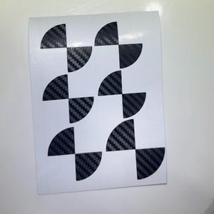 May include: A white rectangular sheet featuring six individual adhesive decals. Each decal consists of two dark gray and black carbon fiber patterned semi-circles arranged to form a circular shape with a small gap between the two halves.