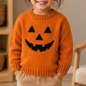 May include: A vibrant orange long-sleeve knit sweater featuring a black jack-o'-lantern face graphic on the front. The sweater has a ribbed crew neckline, ribbed cuffs, and a ribbed hem, paired with light tan corduroy trousers with front pockets.