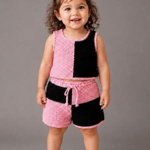 May include: A two-piece crochet set consisting of a sleeveless tank top and matching shorts. The garments feature a colour-blocked design with alternating panels of solid pink and black yarn. The shorts include a drawstring waist detail.