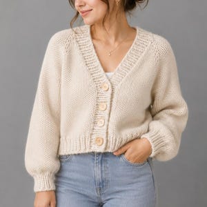 May include: A cream-colored, chunky knit cardigan featuring a V-neckline, long sleeves with ribbed cuffs, and a button-down front with four large, light-colored circular buttons. The garment has a cropped length and a ribbed hemline.