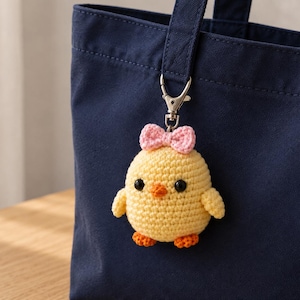 May include: A yellow crocheted chick keychain featuring a small pink bow on its head, black bead eyes, an orange beak, and orange feet. The chick is attached to a silver-toned metal lobster clasp, which is clipped onto a dark navy blue tote bag.