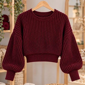 May include: A deep burgundy long-sleeve sweater featuring a thick ribbed knit texture, a rounded crew neckline, and voluminous balloon sleeves with fitted ribbed cuffs. The garment is cropped at the waist with a wide ribbed hemline.