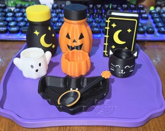 May include: A collection of Halloween-themed items including a black jar with a yellow lid, an orange pumpkin jar, a black book with a yellow moon, a white ghost cup, an orange pumpkin cup, a black cat cup, and a black bat-shaped tray with a spoon.