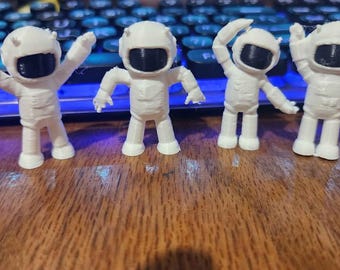 May include: Four small, white, 3D-printed astronaut figurines standing in a row. Each figure features a smooth white suit with a dark, opaque black visor covering the face area. The astronauts are posed with arms in various positions, including raised and extended.