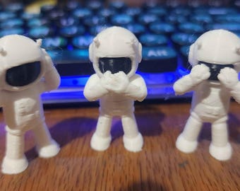May include: Three small white astronaut figurines standing in a row. The left figure covers its ears, the center figure covers its mouth, and the right figure covers its eyes. Each astronaut has a smooth white suit and a black visor.