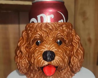 May include: A brown, curly-haired dog-shaped beverage can cooler featuring a black nose, black eyes, and a bright red tongue. A red Dr. Pepper soda can is inserted into the top of the holder, which is resting on a white circular base.