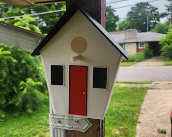 May include: A white birdhouse with a black roof, a central red door, two black windows, and a circular opening above a small wooden perch. The structure is mounted on a wooden post, featuring a clean, geometric design with high-contrast colors.