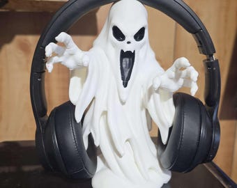 May include: A white 3D-printed ghost figurine with a screaming expression and outstretched arms, wearing a pair of black over-ear headphones. The ghost has hollow black eyes and a wide open mouth, serving as a stand for the black audio headset.