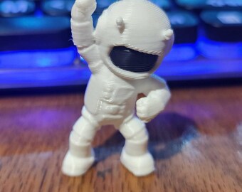 May include: A small, white, 3D-printed figurine of an astronaut in a dynamic pose with one arm raised. The figure features a smooth, rounded helmet with a dark, horizontal visor and a textured suit with visible layer lines from the printing process.