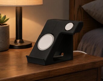 May include: A matte black charging stand featuring a slanted front panel with a circular white magnetic charging pad and a smaller circular white charging disc on the side arm, designed to hold electronic devices securely in place.