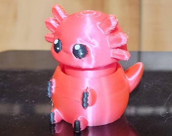 May include: A small, bright pink 3D-printed axolotl figurine with a rounded body, a smooth head, and external gills on both sides. The creature features large, black, circular eyes with white highlights and small black-tipped feet.