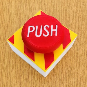 May include: A square-based push button featuring a bright red circular top with the word PUSH printed in white capital letters. The base is yellow with diagonal red stripes on the sides and a white bottom edge, creating a high-contrast design.