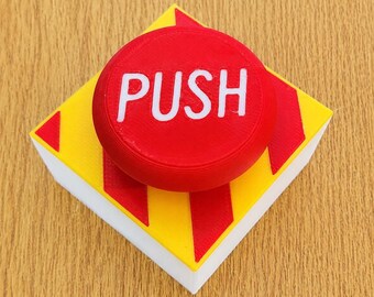 May include: A square-based push button featuring a bright red circular top with the word PUSH printed in white capital letters. The base is yellow with diagonal red stripes on the sides and a white bottom edge, creating a high-contrast design.