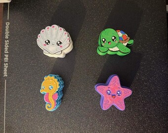 May include: Four small, colorful plastic charms featuring cartoon-style sea creatures: a white seashell, a green turtle, a yellow seahorse, and a pink starfish. Each charm has large, expressive eyes and a smiling mouth, designed as decorative accessories.