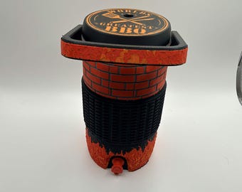May include: A cylindrical beverage cooler with a black lid featuring the text "WORLD'S GREATEST BBQ" and a spatula icon. The body has a red brick pattern, a black textured middle section, and a base decorated with orange and red flame graphics.