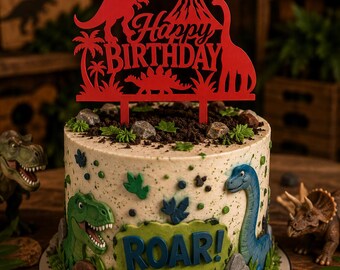 May include: A round cake decorated with green dinosaur figures, rock-like accents, and a green plaque with the text ROAR!. A red cake topper features silhouettes of a T-Rex, a volcano, a long-necked dinosaur, palm trees, and the text Happy BIRTHDAY.