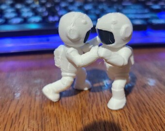 May include: Two small white 3D printed astronaut figurines standing face to face in a boxing stance. Each astronaut wears a white space suit with a smooth finish and a dark black visor covering the face area. The figures are positioned upright.