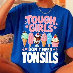 May include: A royal blue t-shirt featuring a graphic print with the text TOUGH GIRLS DON'T NEED TONSILS in pink and white. The design includes illustrations of a mint ice cream cone, a pink popsicle, a chocolate sundae, a soft serve, a blue popsicle, and a cupcake.