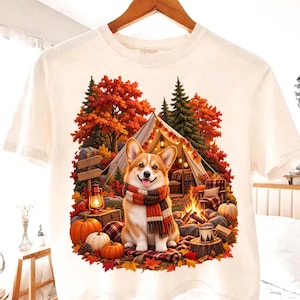 May include: A white short-sleeve t-shirt featuring a graphic of a Corgi wearing a striped scarf sitting in front of a camping tent. The scene includes a campfire, orange and red autumn trees, pumpkins, a lantern, and a wooden signpost.