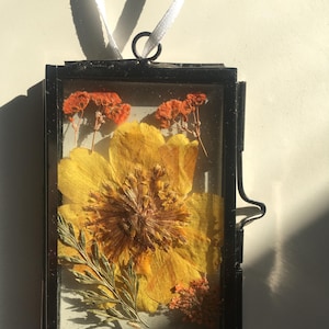 Peut inclure: Un cadre en verre rectangulaire avec un fin bord en métal noir contenant des fleurs séchées pressées. La composition présente une grande fleur jaune au centre, entourée de petites fleurs orange et de feuillage vert. Un ruban blanc est fixé au sommet.