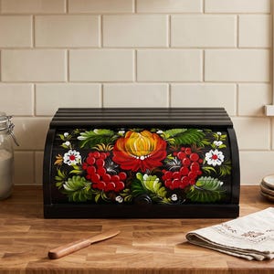 Könnte beinhalten: Ein schwarzer Brotkasten aus Holz mit einem Rolldeckel, der mit einem handgemalten Blumenmuster verziert ist. Das Motiv zeigt eine große, orange-gelbe Blume in der Mitte, umgeben von roten Beeren, weißen Blüten und grünen Blättern auf schwarzem Grund.