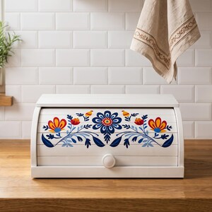 May include: A white wooden bread box with a roll-top lid featuring a colorful floral pattern in shades of blue, red, and yellow. The design includes symmetrical stylized flowers and leaves across the front panel, which also has a small round knob.