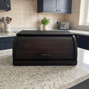 May include: A dark brown wooden bread box with a roll-top lid and a rectangular handle. The bread box features a horizontal slat design on the top and a smooth, curved front panel, showcasing the natural wood grain texture throughout the piece.