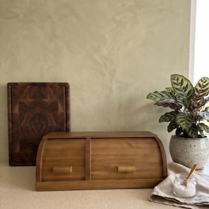 May include: A rectangular wooden bread box with a curved roll-top lid and two small horizontal handles. The bread box is crafted from light brown wood and features a smooth finish, providing a functional storage solution for kitchen countertops.