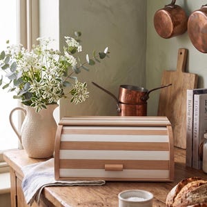 Puede incluir: Una panera rectangular con tapa enrollable que presenta rayas horizontales alternas de madera clara natural y color blanco. La panera tiene un pequeño tirador de madera centrado en el panel frontal y un acabado liso y limpio.