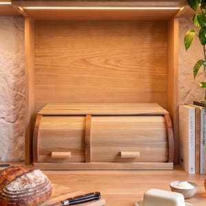 May include: A light-colored wooden bread box with a dual-compartment roll-top design. The unit features two horizontal sliding doors, each equipped with a small rectangular handle, and is constructed from smooth, natural-toned wood planks.